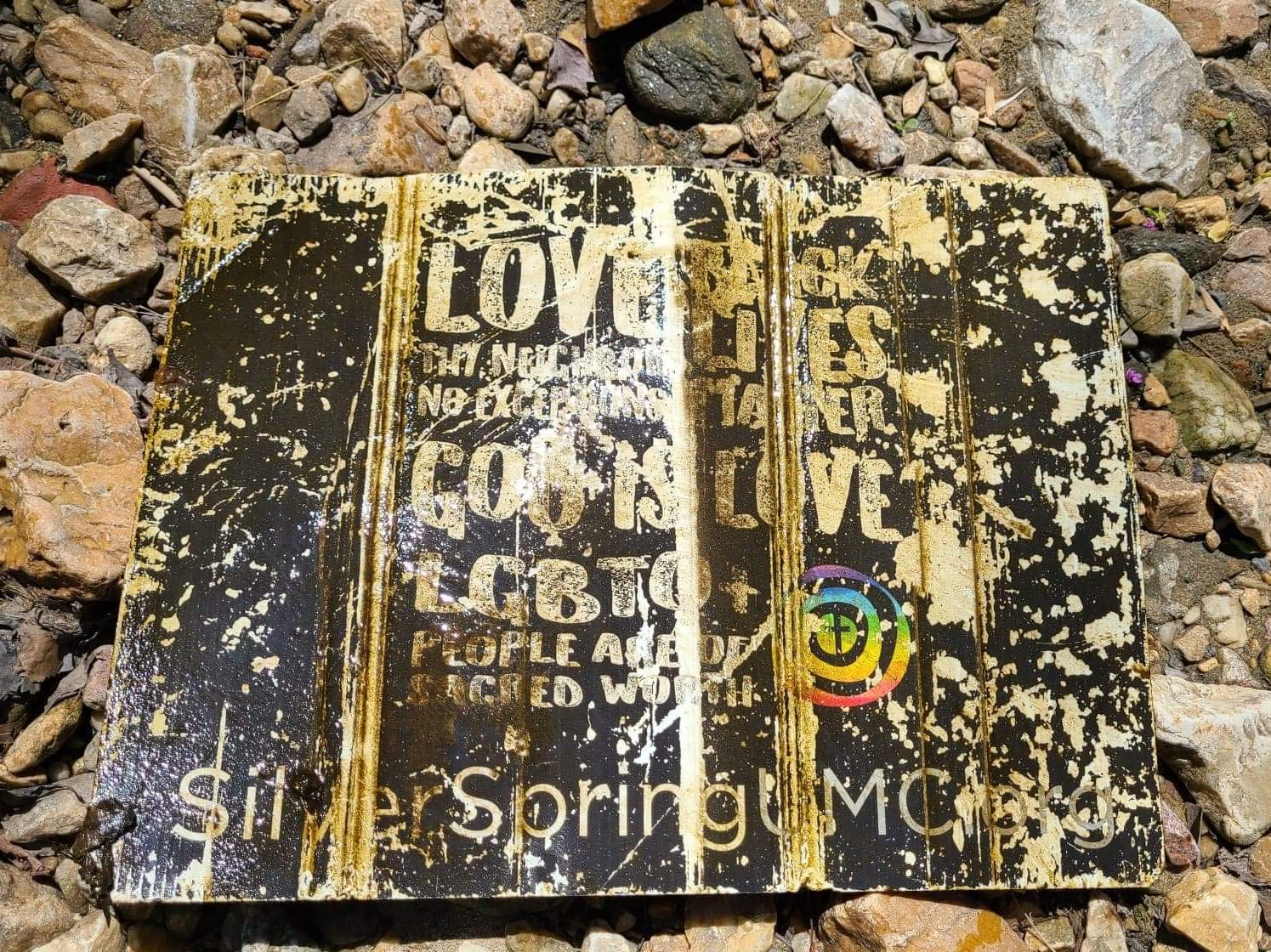 A faded, wet, beaten-up sign saying "Love thy neighbor, no exceptions. Black lives matter. God is love. LGBTQ+ people are of sacred worth."