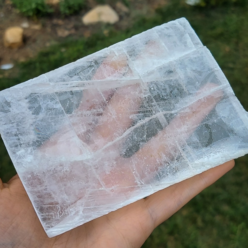 A roughly rectangular selenite plate balanced on a hand. The hand is visible through the selenite, save for a few areas where healed fractures or inclusions give the stone a slightly cloudy appearance. 