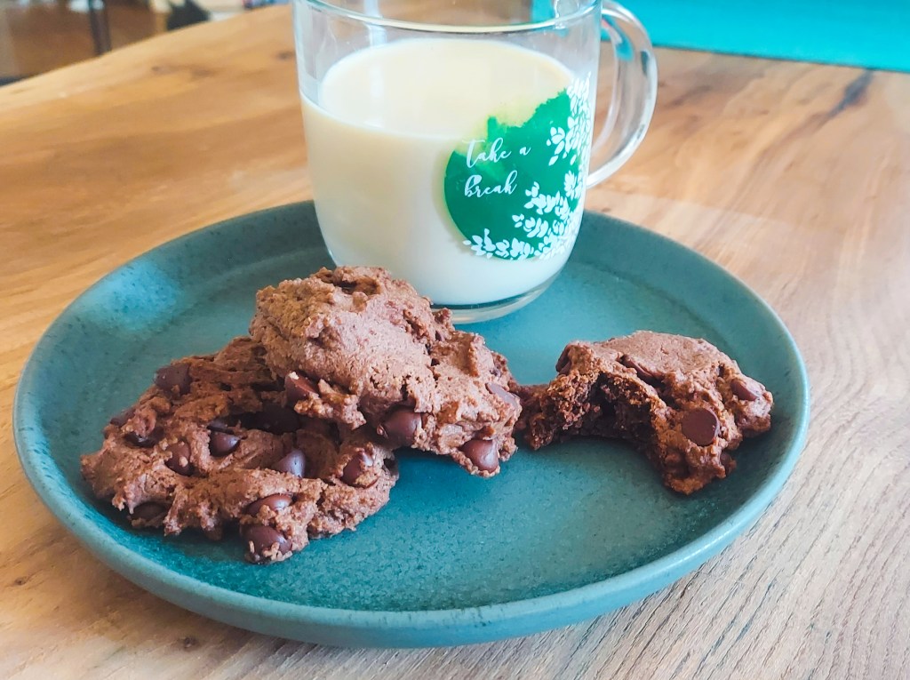 A glass mug of pea milk and three double chocolate cookies, on a green stoneware plate. 