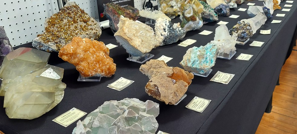 A long table of rare, high-end mineral specimens. There are massive, brightly colored calcites, chalcopyrite, and minerals of every description, in every hue.
