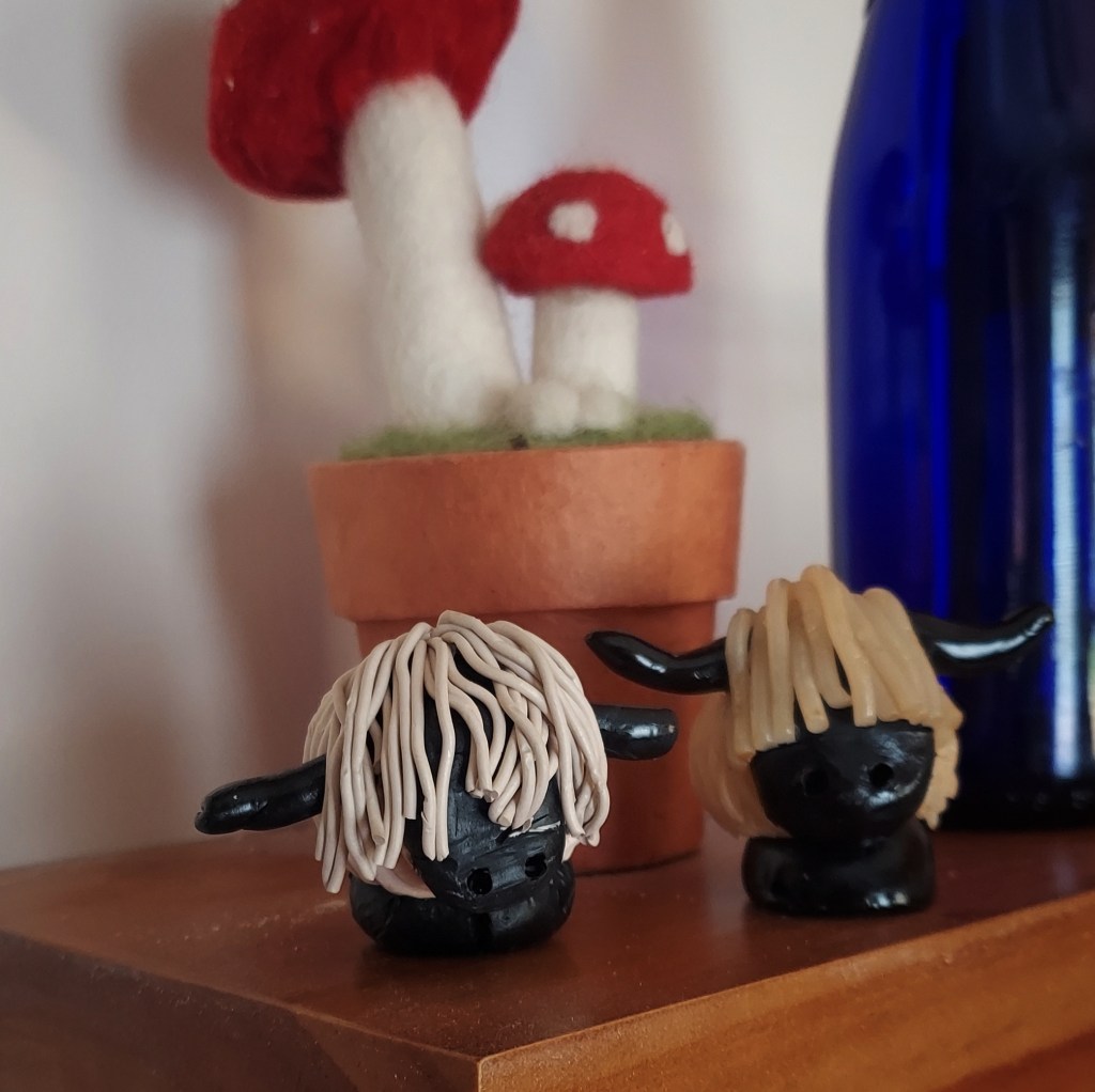 A pair of small, polymer clay sheep in front of a blue glass bottle and a needlefelted Amanita muscaria.