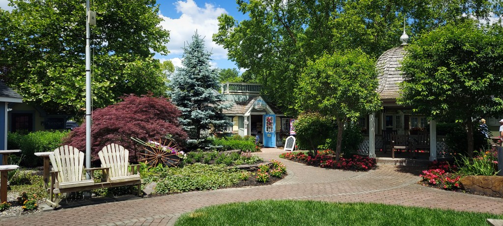 Another view of Woodland Village, showing a gazebo, brick pathways lined with flowers and trees, an old-fashioned wheelbarrow full of plants, a set of wooden Adirondak chairs, and a shop in the background.