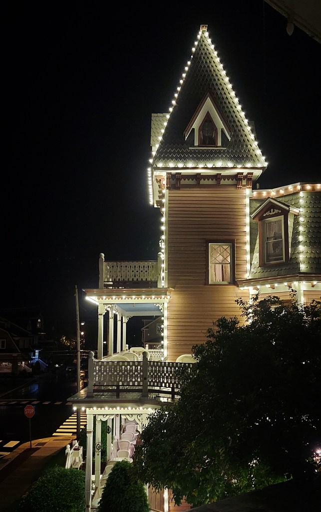 A view of a pink Victorian house at night. It has "gingerbread" details and a scalloped roof and is lit with strings of cafe lights.