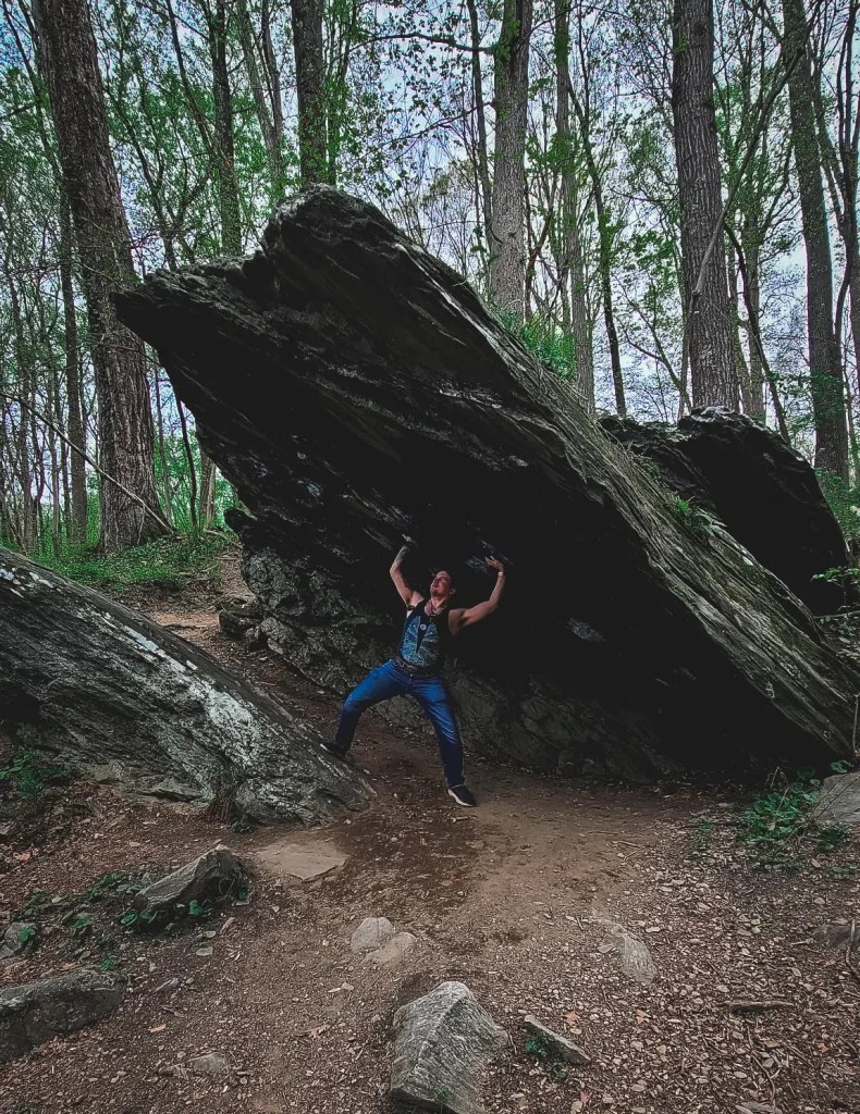 My Handsome Assistant, pretending to hold up the rock shelter. The "shelter" itself is a very large, flat boulder jutting up at an angle, creating a kind of natural awning over a bare patch of soil.