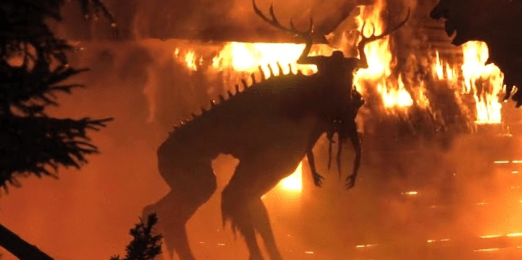 A large monster, silhouetted against a burning building. The monster appears to be a large, quasi-skeletal elk with a head made up of an upside-down human corpse. The arms hang down like mandibles, and the legs rise up and branch into antlers. A pair of tiny, glowing eyes are visible where the corpse's head would be. 