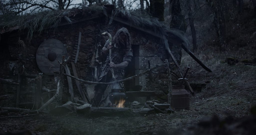 The Father, outside of his small house. He uses a large stick to pound something boiling in an iron cauldron.